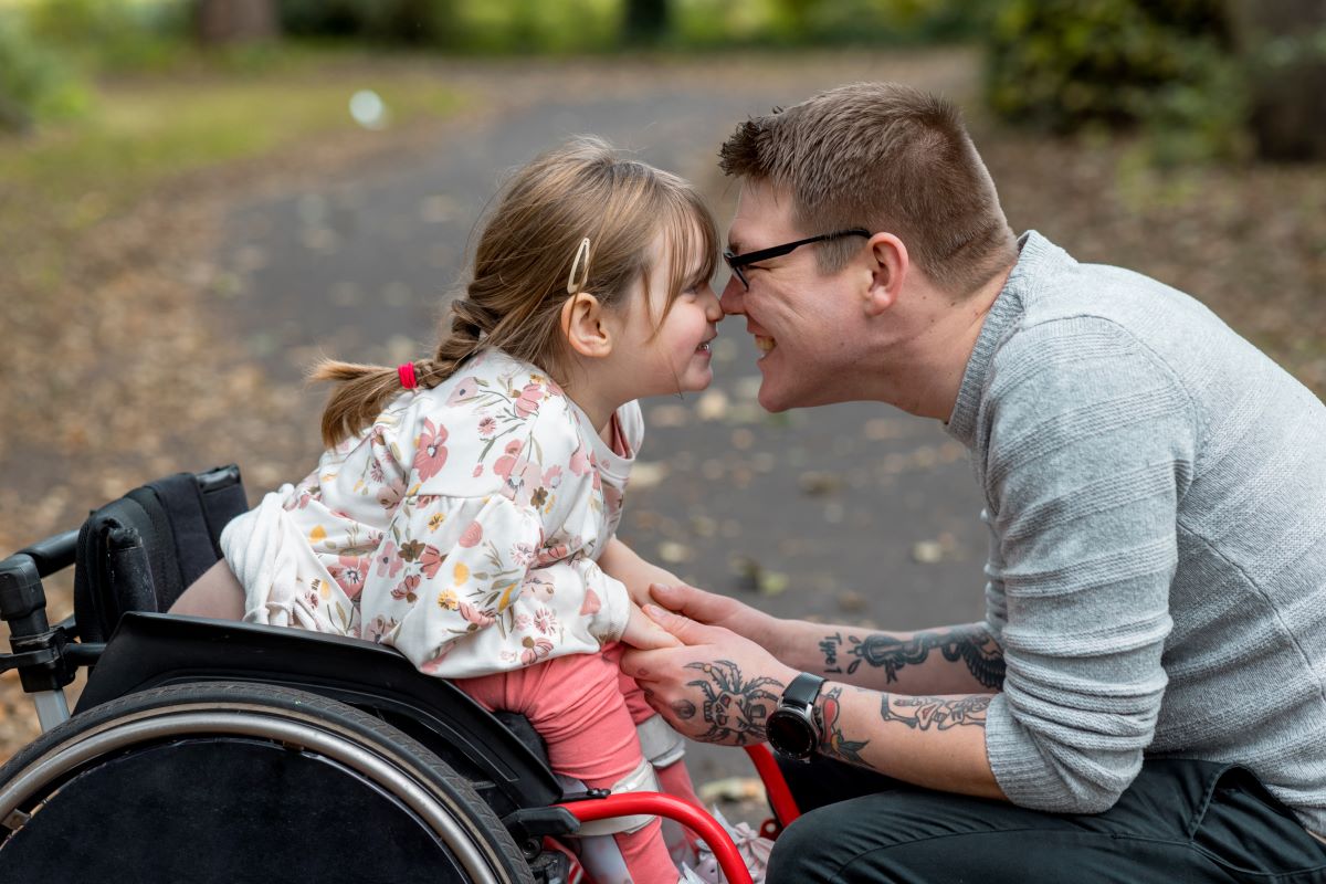 Pediatrics-breathing-disorders-treatment Older person in a park is crouched in order to press their nose against the nose of a child in a wheelchair opposite of them. Both people are smiling.