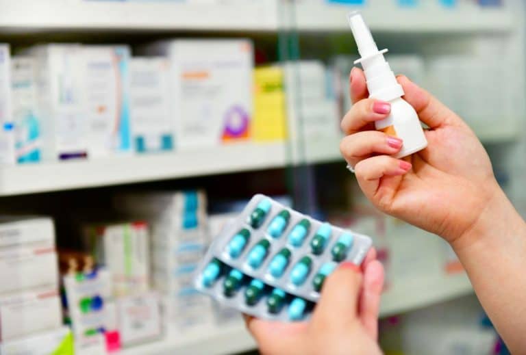 Packages of Allergy Medication A blurry background of a drugstore shelf. Two hands are in frame and in focus. One hand holds two packets of green and blue pills while the other hand holds a nasal spray bottle.