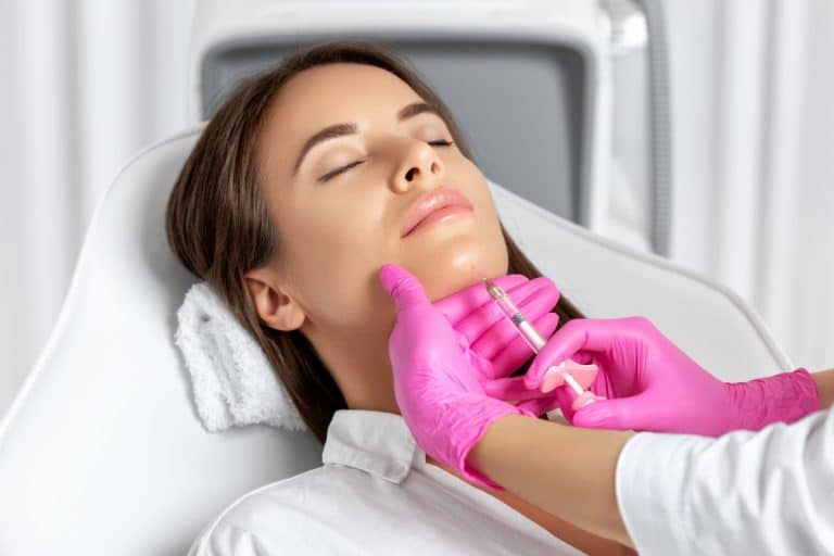 Facial Implant Patient Woman with eyes closed on a medical table with a towel beneath her head. Her eyes are closed and a pair of orange gloved hands coming in from the right of the frame inject a solution into her chin.